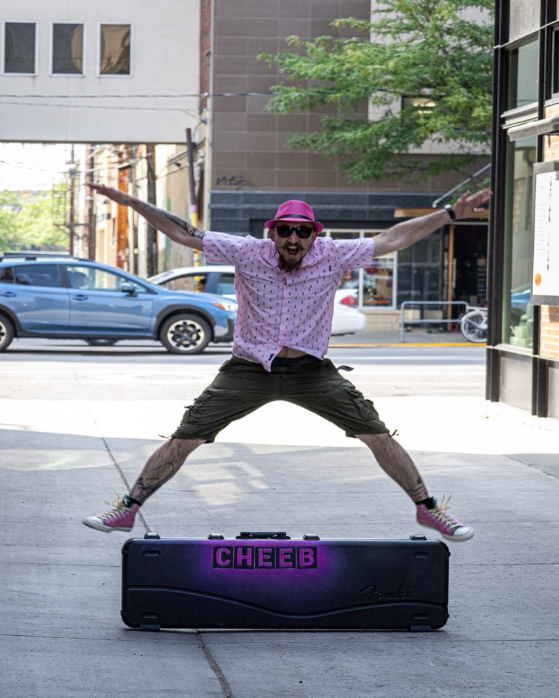 Cheeb mid-jump on a downtown sidewalk, arms and legs flung wide, clearing the CHEEB flight case below him