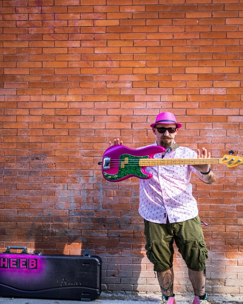 Cheeb holding his pink Precision bass flat out toward the camera in front of a red brick wall