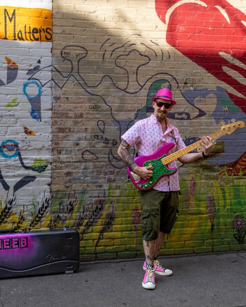 Cheeb playing bass beside a painted mural reading &ldquo;Your Tomorrow Matters&rdquo;, his CHEEB case on the ground