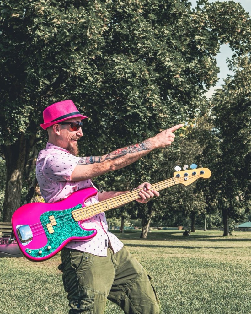 Cheeb pointing off to one side mid-song on a tree-lined park lawn, bass slung across him