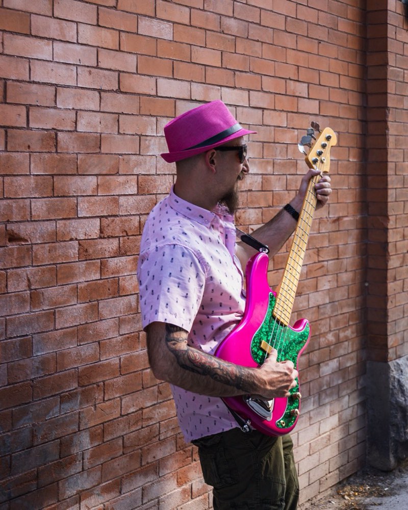 Cheeb in profile close to a red brick wall, pink fedora and sunglasses, right hand on the strings